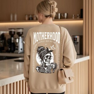 Pu&ograve; includere: Felpa beige con la scritta "MOTHERHOOD" stampata sul retro. La grafica presenta uno scheletro con una bandana che tiene una tazza. Il testo sulla felpa recita "Some days I rock it, some days it rocks me, anyways we're rockin".