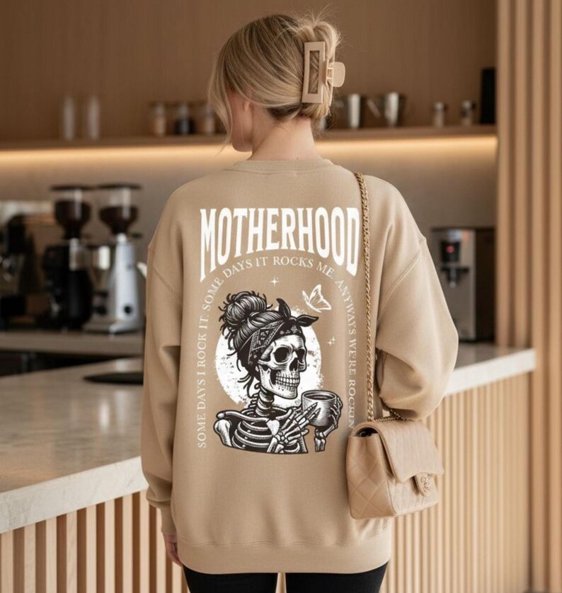 Pu&ograve; includere: Felpa beige con la scritta "MOTHERHOOD" stampata sul retro. La grafica presenta uno scheletro con una bandana che tiene una tazza. Il testo sulla felpa recita "Some days I rock it, some days it rocks me, anyways we're rockin".