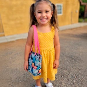 May include: A young child wearing a yellow polka-dot dress and white sneakers, holding a tote bag with a cat pattern. The bag has a pink strap. The child is smiling, with pigtails, and standing on a gravel surface.