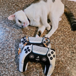 May include: Two plush video game controller pillows featuring a white and black design with iconic geometric symbols. One pillow displays a pattern of cat paws, while the other features a central PlayStation logo and a grid of controller button icons.