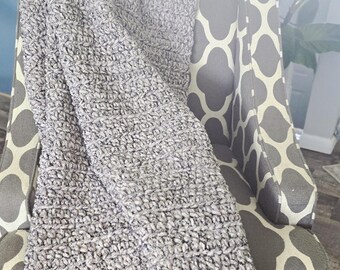 May include: A grey, textured blanket draped over a patterned armchair. The blanket appears soft and plush, with a close-knit weave. The chair has a grey and white geometric design, with wooden legs. The background includes greenery.