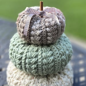 Puede incluir: Una pila de tres calabazas tejidas a mano en colores otoñales. La calabaza superior es marrón con un tallo de canela y una cinta a cuadros. La calabaza del medio es verde y la inferior es de color crema.