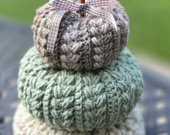 May include: A stack of three handmade, knitted pumpkins in fall colors. The top pumpkin is brown with a cinnamon stick stem and a checkered ribbon. The middle pumpkin is green, and the bottom pumpkin is cream-colored.