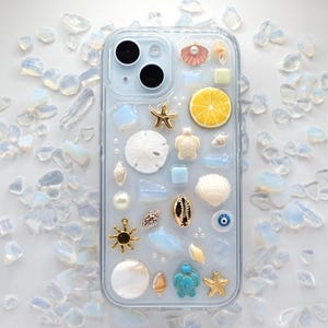 May include: A clear phone case with a light blue background and various sea-themed decorations, including seashells, a starfish, a turtle, a lemon slice, and a blue evil eye charm.
