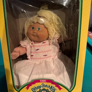 May include: A 1985 Cabbage Patch Kids doll in a white dress with pink and red trim. The doll has blonde hair and blue eyes. The doll is in its original box, which is yellow with green trim. The box has the text "1985 Cabbage Patch Kids" and "I'm unique. I'm adoptable!" on it.
