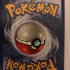 Puede incluir: Primer plano de una carta de intercambio de Pokemon con un borde azul y un fondo gris en espiral. El logotipo de Pokemon est&aacute; en la esquina superior derecha y el logotipo de Pokeball est&aacute; en la esquina inferior izquierda. La carta presenta una Pokeball con un dise&ntilde;o rojo y blanco.