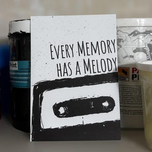 May include: A white card with the text "EVERY MEMORY HAS A MELODY" in black, above a black and white cassette tape illustration. The card is set against a neutral background with paint containers.