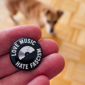 Könnte beinhalten: Ein runder, schwarzer Ansteckbutton mit weißem Text, der LOVE MUSIC HATE FASCISM lautet, und einer zentralen Grafik einer weißen Schallplatte. Das Design ist auf einer glatten, runden Oberfläche mit klarer Schrift und hohem Kontrast gedruckt.