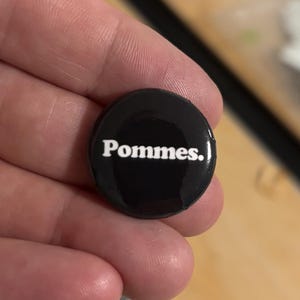May include: A small, round, black pin-back button featuring the word "Pommes." printed in a bold, white, sans-serif font across the center. The surface of the button is smooth and glossy, reflecting light across its circular, dark-colored face.