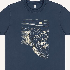 Pu&ograve; includere: T-shirt blu navy con un disegno di paesaggio color crema. La grafica raffigura una costa con rocce, acqua e un sole o una luna. La maglietta ha un girocollo e maniche corte.