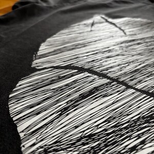 May include: Black t-shirt with a large, abstract white design. The design features a series of parallel, scribbled lines, creating a textured effect. The shirt is made of a soft material and is slightly wrinkled.