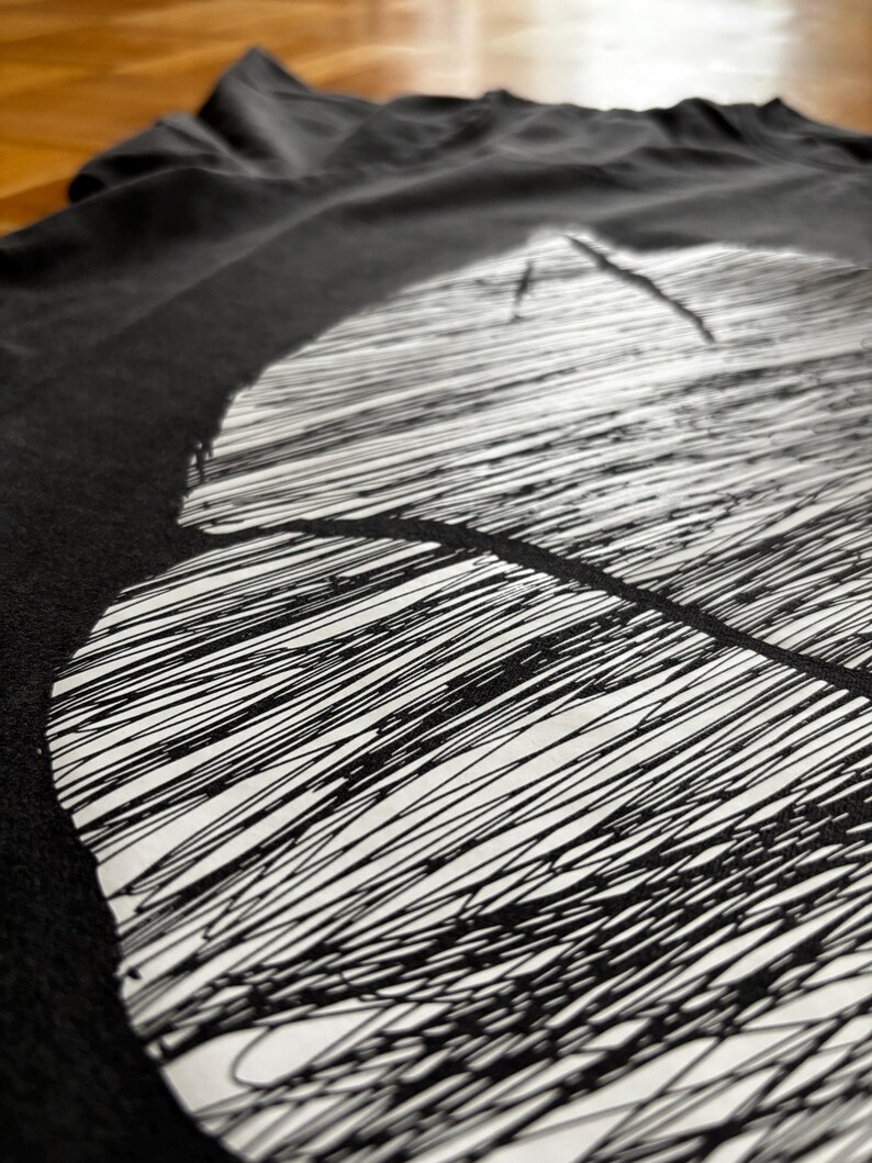 May include: Black t-shirt with a large, abstract white design. The design features a series of parallel, scribbled lines, creating a textured effect. The shirt is made of a soft material and is slightly wrinkled.