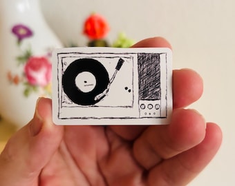 May include: A rectangular sticker featuring a black and white ink sketch of a record player. The design includes a circular vinyl record, a tonearm, and a speaker section with three circular knobs, all rendered in a hand-drawn, sketchy style.