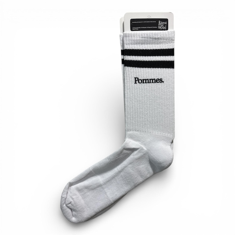 May include: A white crew sock featuring two horizontal black stripes at the top cuff and the word Pommes. printed in black text below the stripes. The sock is attached to a white cardboard header card with small black text at the top.