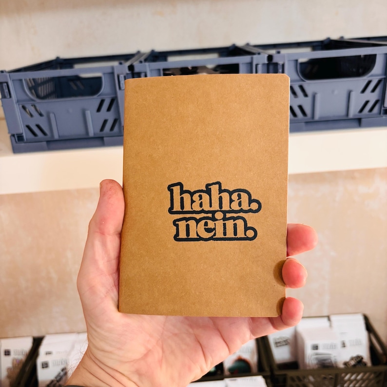 May include: A tan rectangular notebook with the black text "haha. nein." printed in the center. The notebook has a smooth, matte cover and is held in front of blue plastic storage crates. The design features bold, rounded, lowercase lettering.