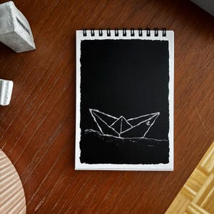 Könnte beinhalten: Ein spiralgebundenes Notizbuch mit einer schwarzen Seite, die eine weiße Strichzeichnung eines Papierbootes auf Wellen zeigt. Das Notizbuch liegt auf einer Holzoberfläche, im Hintergrund befindet sich ein grauer Betongegenstand.