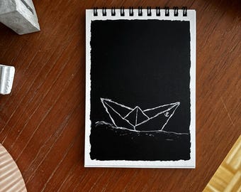 May include: A spiral-bound notepad with a black page featuring a white line drawing of a paper boat sailing on waves. The notepad is on a wooden surface, with a gray concrete object in the background.