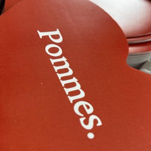 May include: Close-up of several heart-shaped coasters. The coasters are a vibrant red with the word "Pommes" printed in white. The coasters are stacked, with some visible in the background.