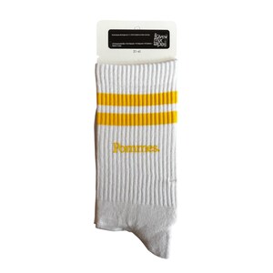 May include: A pair of white crew socks featuring two horizontal yellow stripes near the top cuff and the word Pommes embroidered in yellow thread on the side. The socks are attached to a white cardboard header card with the text i love mix tapes.