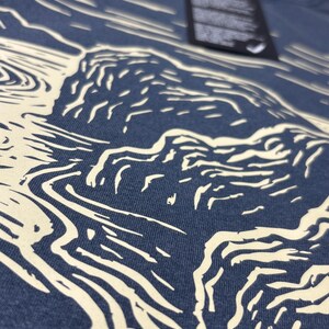 Pu&ograve; includere: Primo piano di una maglietta blu navy con una grafica color crema raffigurante un paesaggio con acqua e scogliere. Il design &egrave; un'illustrazione stilizzata. Un'etichetta nera &egrave; appesa alla maglietta.