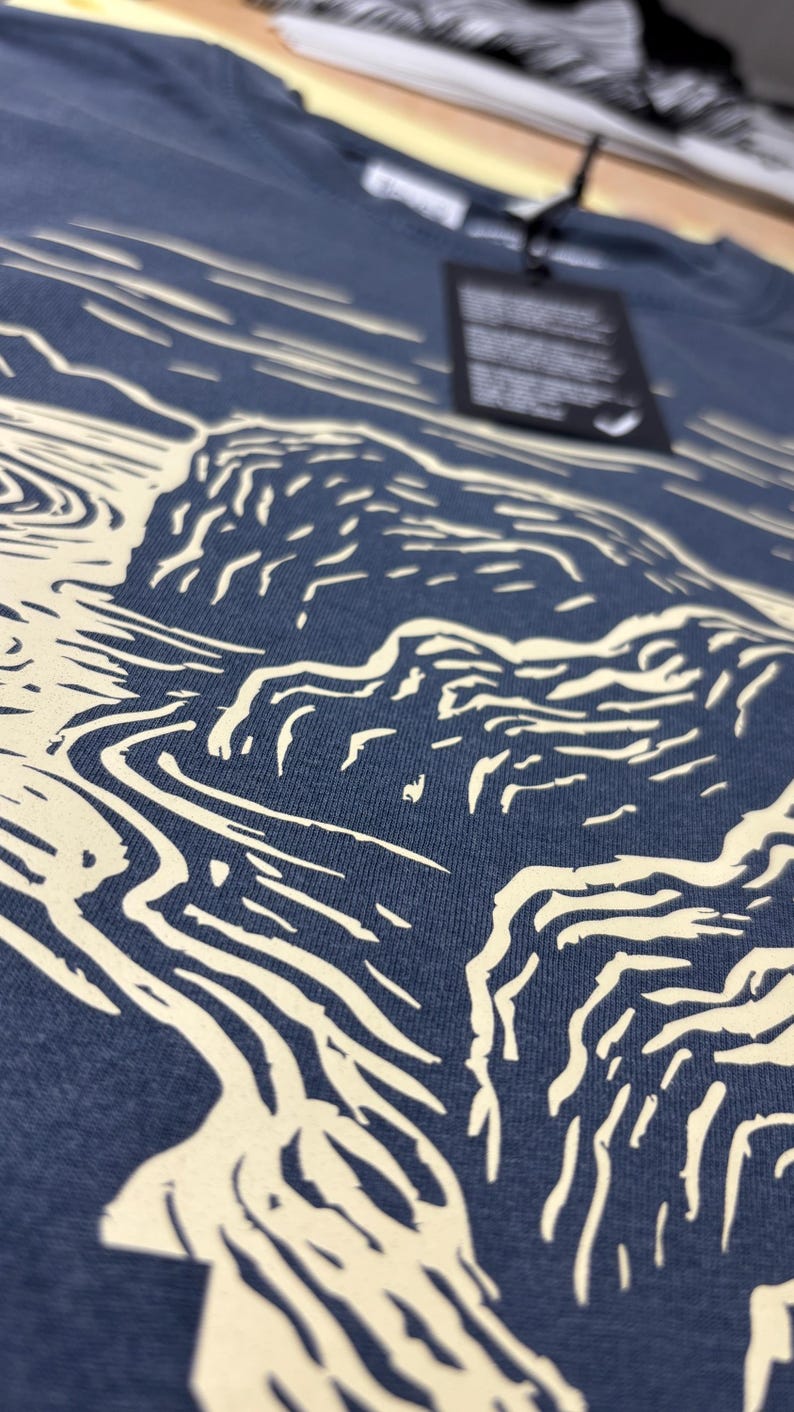 Pu&ograve; includere: Primo piano di una maglietta blu navy con una grafica color crema raffigurante un paesaggio con acqua e scogliere. Il design &egrave; un'illustrazione stilizzata. Un'etichetta nera &egrave; appesa alla maglietta.