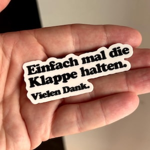 Könnte beinhalten: Ein weißer, konturgestanzter Aufkleber mit schwarzem Text, der "Einfach mal die Klappe halten. Vielen Dank." lautet. Der Aufkleber hat eine individuelle Form, die dem Verlauf der drei Textzeilen folgt, und verwendet eine fette, serifenlose Schrift.
