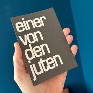 May include: A dark gray card with the words "einer von den juten" in white, held against a blue background. The text is in a bold, sans-serif font.