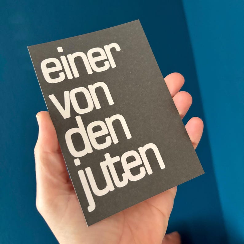 May include: A dark gray card with the words "einer von den juten" in white, held against a blue background. The text is in a bold, sans-serif font.