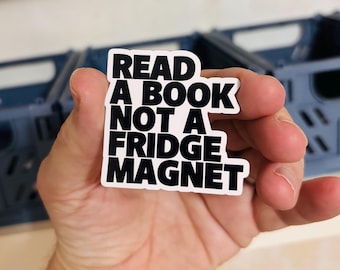 May include: A die-cut sticker with a white background and bold black text that reads: READ A BOOK NOT A FRIDGE MAGNET. The sticker features a clean, sans-serif font and a simple, rectangular-inspired shape with rounded corners.