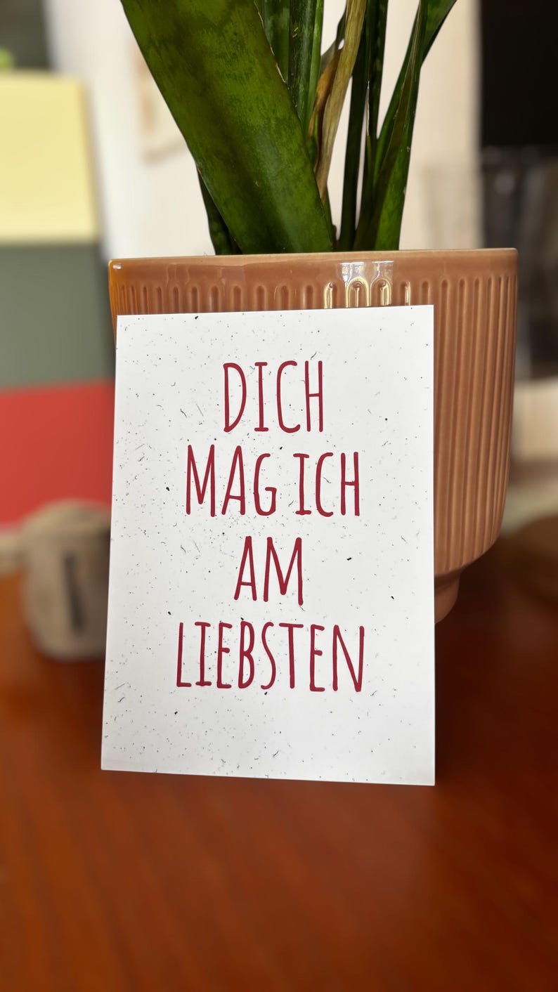 May include: A rectangular white card with a speckled texture featuring the red text DICH MAG ICH AM LIEBSTEN. The card is positioned in front of a ribbed, light brown ceramic planter containing a green plant with long, pointed leaves.