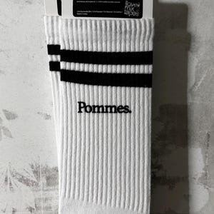 May include: A pair of white ribbed crew socks featuring two horizontal black stripes near the top cuff and the word Pommes. embroidered in black lettering on the side. The socks are attached to a white cardboard retail header card with text.
