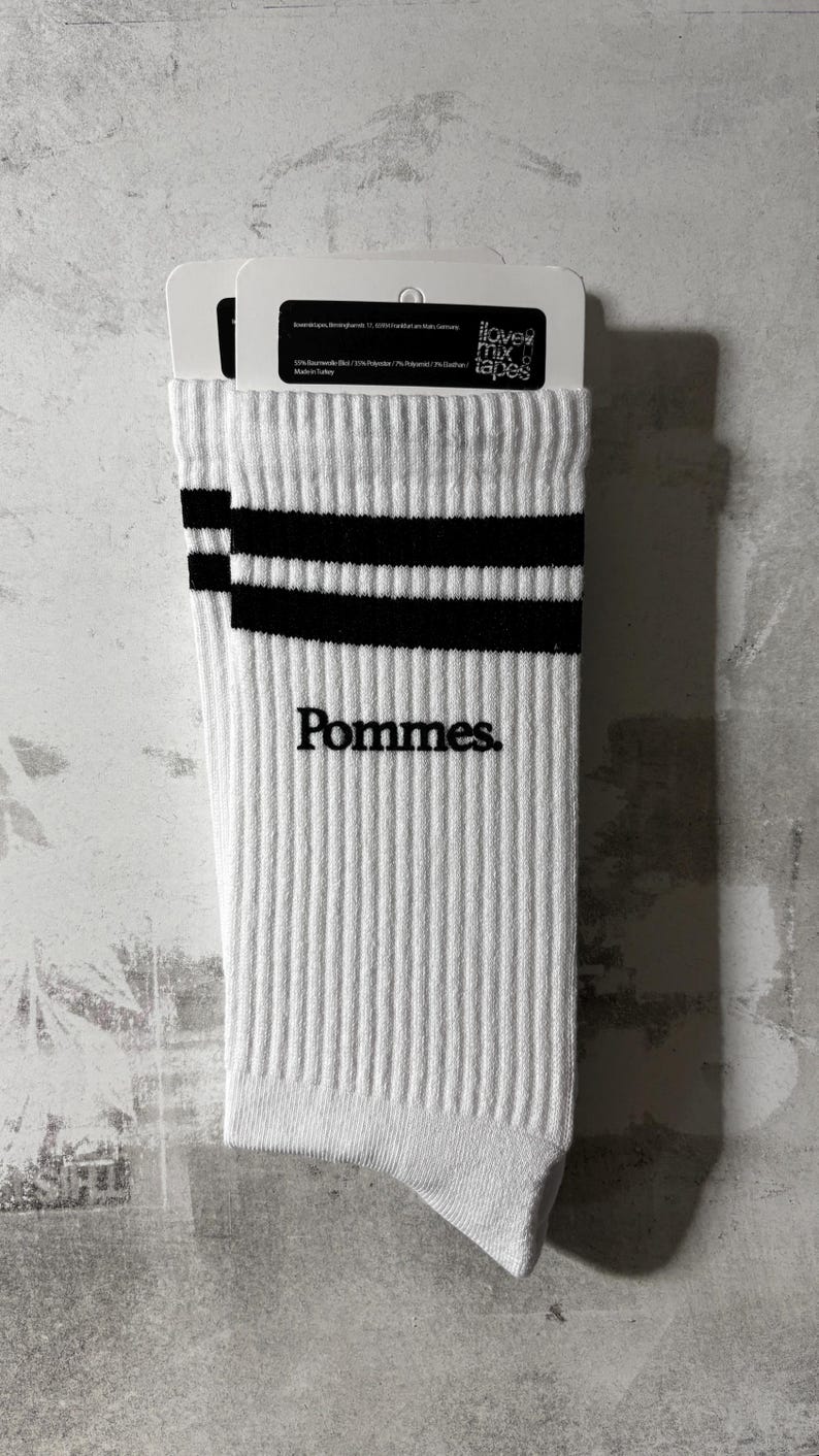 May include: A pair of white ribbed crew socks featuring two horizontal black stripes near the top cuff and the word Pommes. embroidered in black lettering on the side. The socks are attached to a white cardboard retail header card with text.