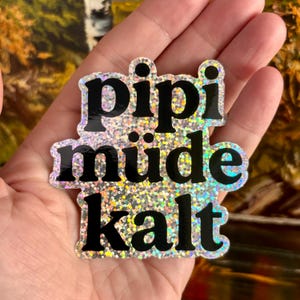 May include: A die-cut sticker with a holographic, silver-toned glitter finish featuring the black text 'pipi', 'müde', and 'kalt' arranged in three stacked lines. The sticker has a reflective, multi-colored surface that catches the light.