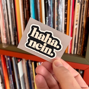 Könnte beinhalten: Ein rechteckiger Aufkleber mit transparentem Hintergrund, der den Text "haha. nein." in einer fetten, cremefarbenen, abgerundeten serifenlosen Schrift mit dickem schwarzem Rand auf einer durchsichtigen Klebefolie zeigt.