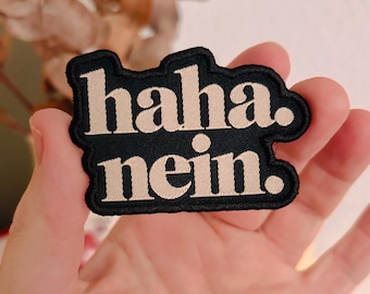 May include: A black embroidered patch with a thick border featuring the text "haha. nein." in a light beige, sans-serif font. The patch has a distinct, irregular shape that follows the contour of the bold, lowercase lettering.
