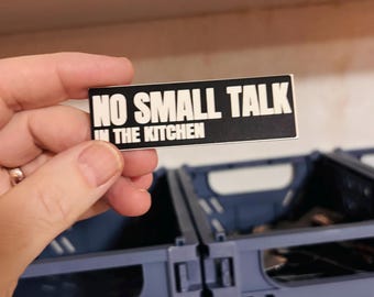 May include: A rectangular black sticker with white text that reads NO SMALL TALK IN THE KITCHEN in a bold, sans-serif font. The sticker has a clean, flat design with sharp corners and a high-contrast color scheme.