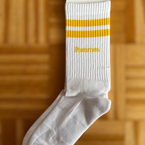 May include: A white crew sock with two horizontal yellow stripes at the top and the word Pommes embroidered in yellow on the side. The sock is attached to a white cardboard tag featuring the text love mix tapes and the size range 37-41.