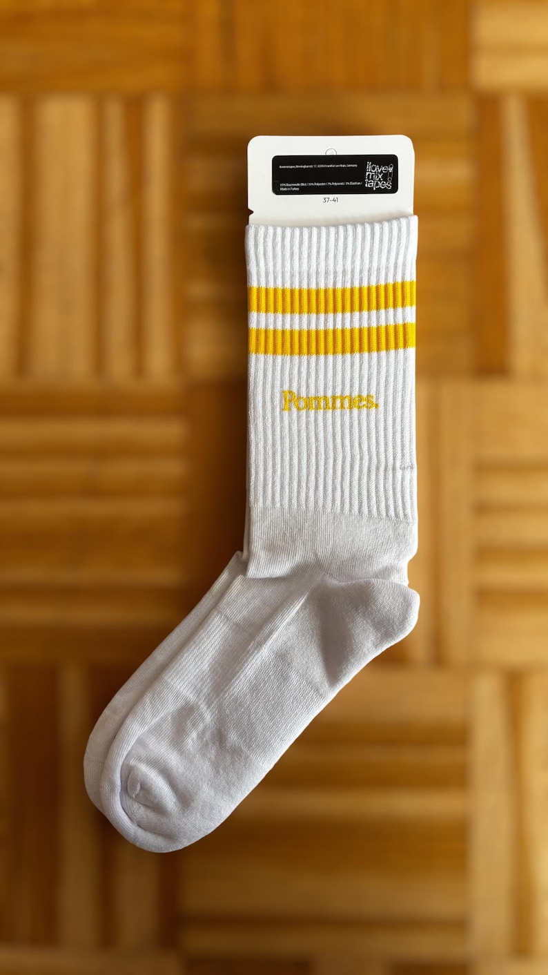 May include: A white crew sock with two horizontal yellow stripes at the top and the word Pommes embroidered in yellow on the side. The sock is attached to a white cardboard tag featuring the text love mix tapes and the size range 37-41.