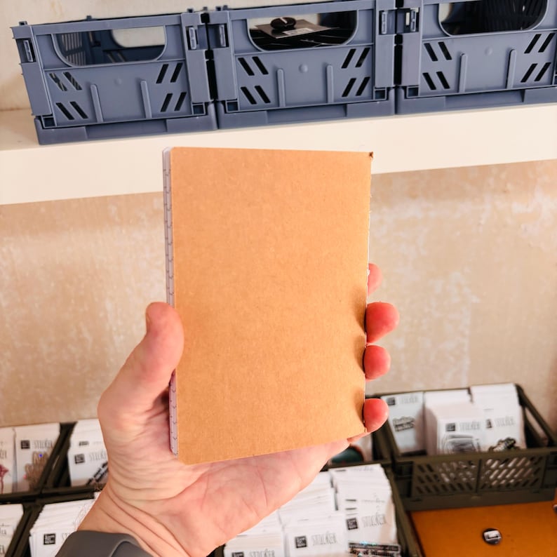 May include: A rectangular notebook with a plain, light brown cardboard cover. The notebook is held upright, displaying its solid tan front surface and a thin white spiral binding visible along the left edge, showcasing a simple and functional design.