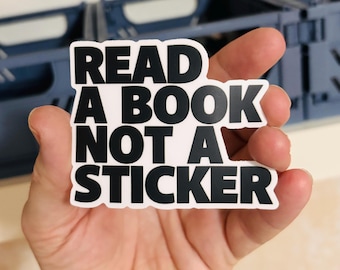 May include: A die-cut sticker with a white background and bold black sans-serif text that reads READ A BOOK NOT A STICKER. The sticker features a clean, blocky font and a distinct white border surrounding the black lettering.