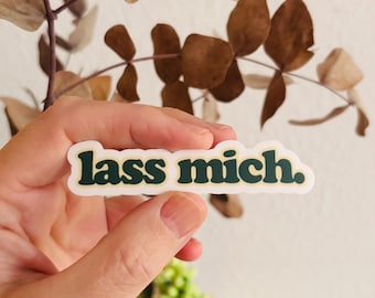May include: A die-cut sticker featuring the text "lass mich." in a dark green, rounded serif font. The lettering is outlined in a thin cream-colored border, creating a clean, high-contrast look for the German phrase displayed on the sticker.