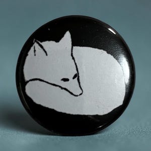 Könnte beinhalten: Ein runder, schwarzer Knopf mit einer weißen Illustration eines sich zusammenrollenden Fuchses. Der Fuchs ist mit einfachen schwarzen Linien dargestellt. Der Knopf hat eine glänzende Oberfläche und ein minimalistisches Design.