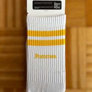 May include: A pair of white crew socks featuring two horizontal yellow stripes near the top and the word Pommes embroidered in yellow on the side. The socks are attached to a white cardboard tag with the text i love mix tapes and size 37-41.