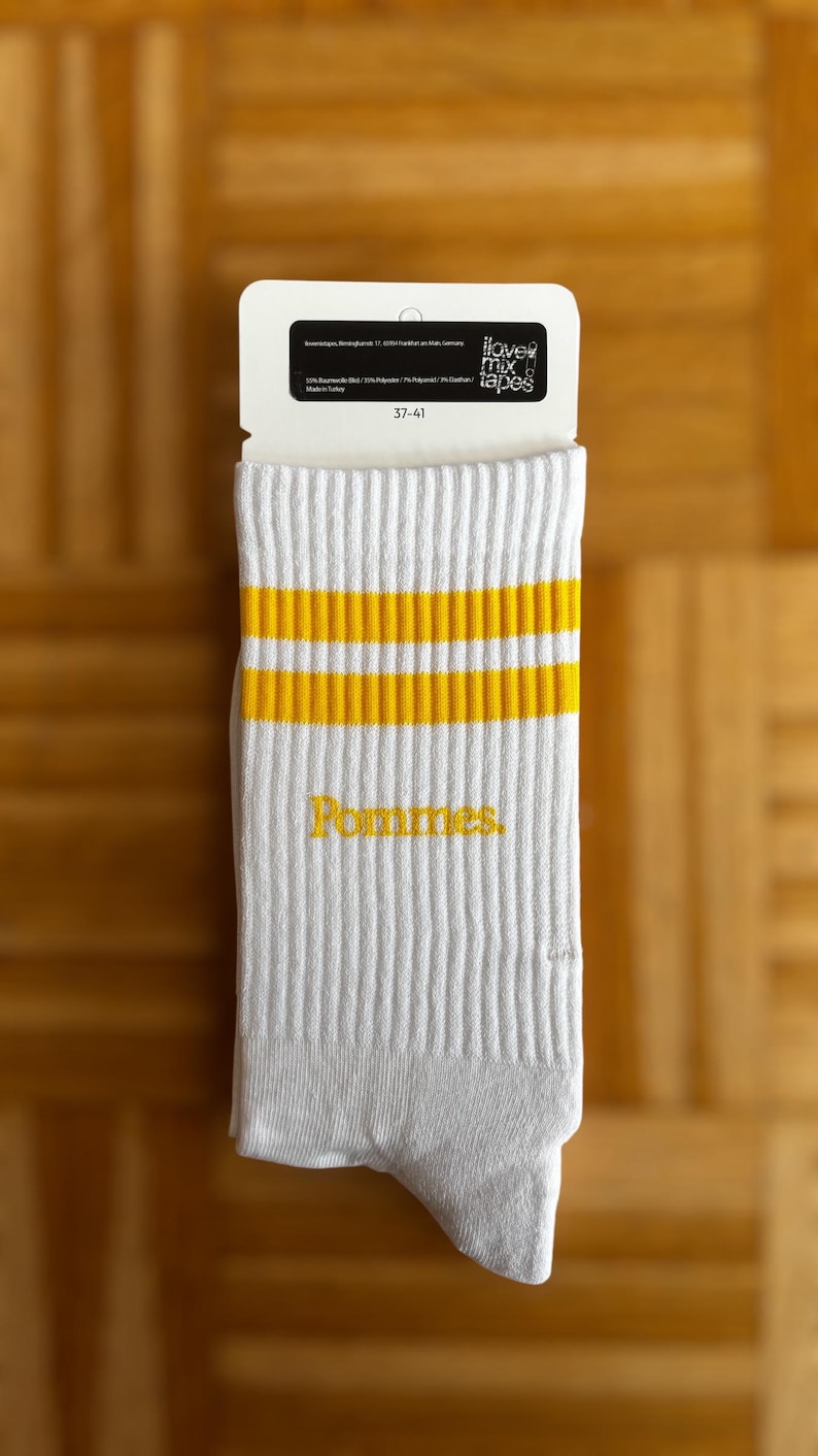 May include: A pair of white crew socks featuring two horizontal yellow stripes near the top and the word Pommes embroidered in yellow on the side. The socks are attached to a white cardboard tag with the text i love mix tapes and size 37-41.