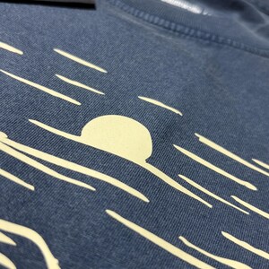 Pu&ograve; includere: Primo piano di una maglietta blu con una grafica color crema di un sole che tramonta sull'oceano. Il design include un grande sole e diverse linee orizzontali. La maglietta ha un'etichetta attaccata con testo.