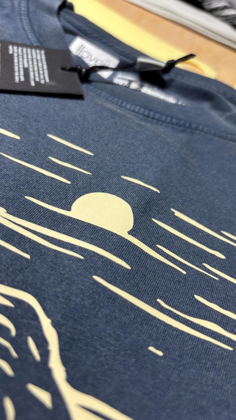 Pu&ograve; includere: Primo piano di una maglietta blu con una grafica color crema di un sole che tramonta sull'oceano. Il design include un grande sole e diverse linee orizzontali. La maglietta ha un'etichetta attaccata con testo.