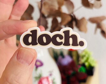 May include: A die-cut sticker featuring the word doch. in a bold, dark brown, rounded lowercase font. The text is outlined in a thick, cream-colored border, creating a clean and simple graphic design element on a white background.