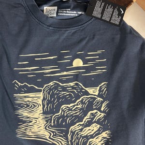 Pu&ograve; includere: T-shirt blu navy con un disegno di paesaggio color crema con sole, acqua e rocce. La maglietta ha uno scollo tondo e un'etichetta. Il marchio "ilovemixtapes" &egrave; visibile sulla maglietta.