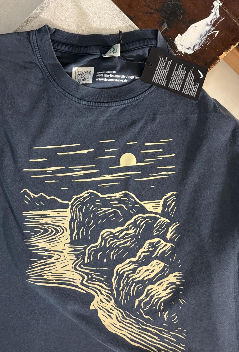 Pu&ograve; includere: T-shirt blu navy con un disegno di paesaggio color crema con sole, acqua e rocce. La maglietta ha uno scollo tondo e un'etichetta. Il marchio "ilovemixtapes" &egrave; visibile sulla maglietta.