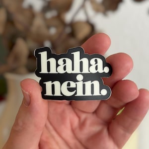 May include: A black and white sticker with the words "haha. nein." in a bold, sans-serif font. The sticker is held in a hand, with a blurred background of dried plants.